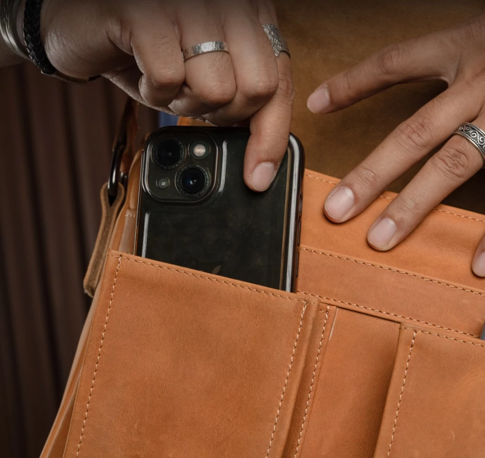 Hand placing smartphone into front pocket of brown leather messenger bag.