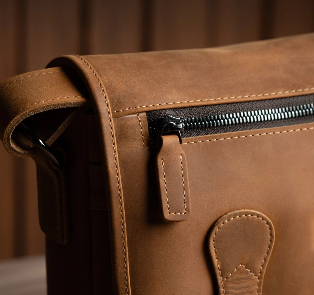 Close-up of brown leather messenger bag showing zipper pocket and stitching detail.